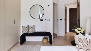 Um quarto com cama, espelho redondo, mesa de cabeceira, cesta e porta que dá para outro cômodo.