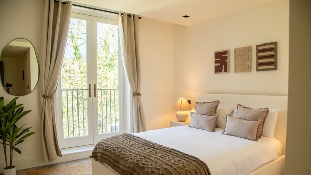 A bedroom with a bed, curtains, a bedside lamp, a mirror, and a potted plant. - The Pembridge 11 (London)