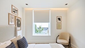 Ein Schlafzimmer mit einem Bett, einem Sessel, einem Fenster mit Jalousien und gerahmten Kunstwerken an der Wand.