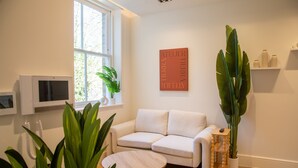 Ein modernes Wohnzimmer mit einem weißen Sofa, einem runden Holztisch, einer großen Küsterpflanze, einem fest montierten Flachbildfernseher und einem Wandregal mit Dekoration.
