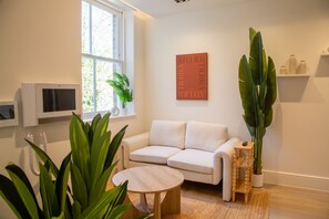 A modern living room with a white sofa, a round wooden coffee table, a large potted plant, a mounted flat-screen TV, and a wall-mounted shelf with decorative items. - The Pembridge 12 (London)