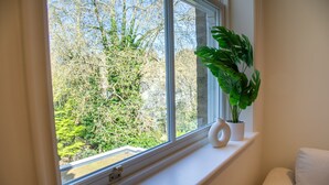 Ein Fenster mit Blick auf Bäume, eine weiße Vase und eine Pflanze auf einem Regal.