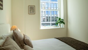 A hotel room with a bed, pillows, a lamp, a window with a plant, and a framed picture on the wall. - Pembridge Residences - Flat 3 (London)