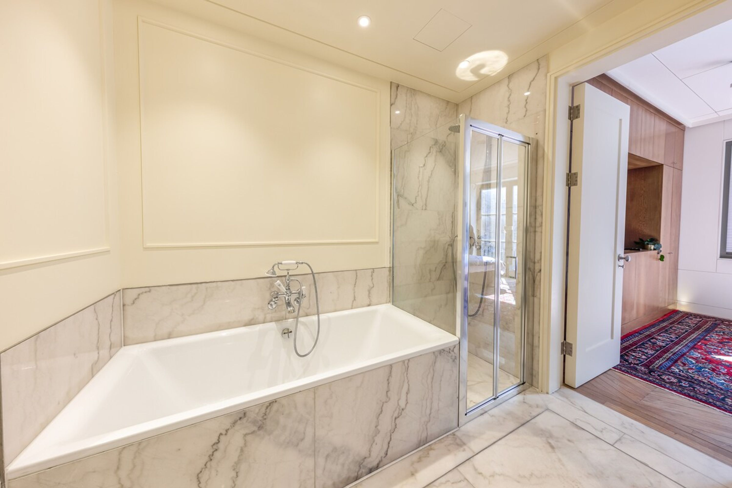 A modern bathroom with a marble bathtub, glass shower enclosure, and a wooden floor.