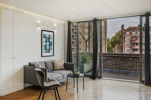 Ein modernes Wohnzimmer mit einem grauen Sofa, einem kleinen Tisch mit einer Pflanze und einem großen Fenster mit Blick auf rote Backsteingebäude.