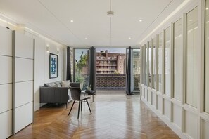 Ein modernes Interieur mit Fischgrätparkett, einem grauen Sofa, einem kleinen Tisch und Blick auf ein Backsteingebäude durch große Fenster.