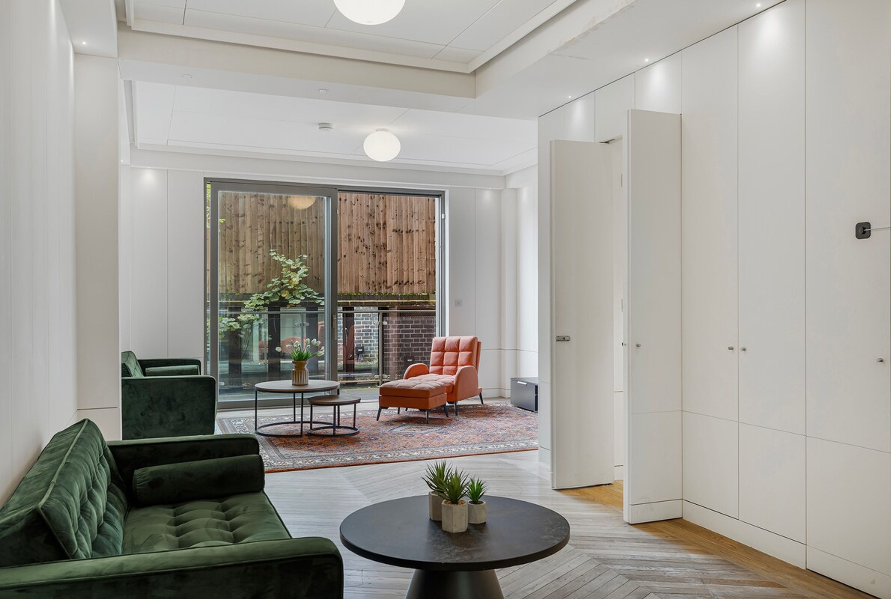 A modern interior with a green sofa, an orange armchair, a round table, and a large window with a view of a brick wall.