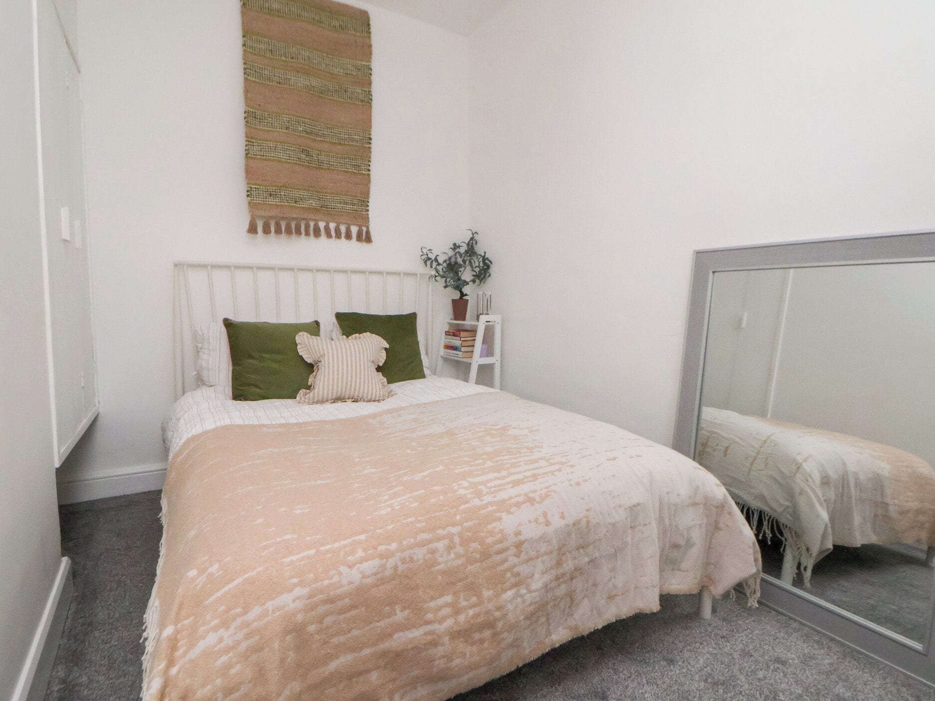 A bedroom with a bed, a mirror, a plant, and a wall-mounted tapestry.