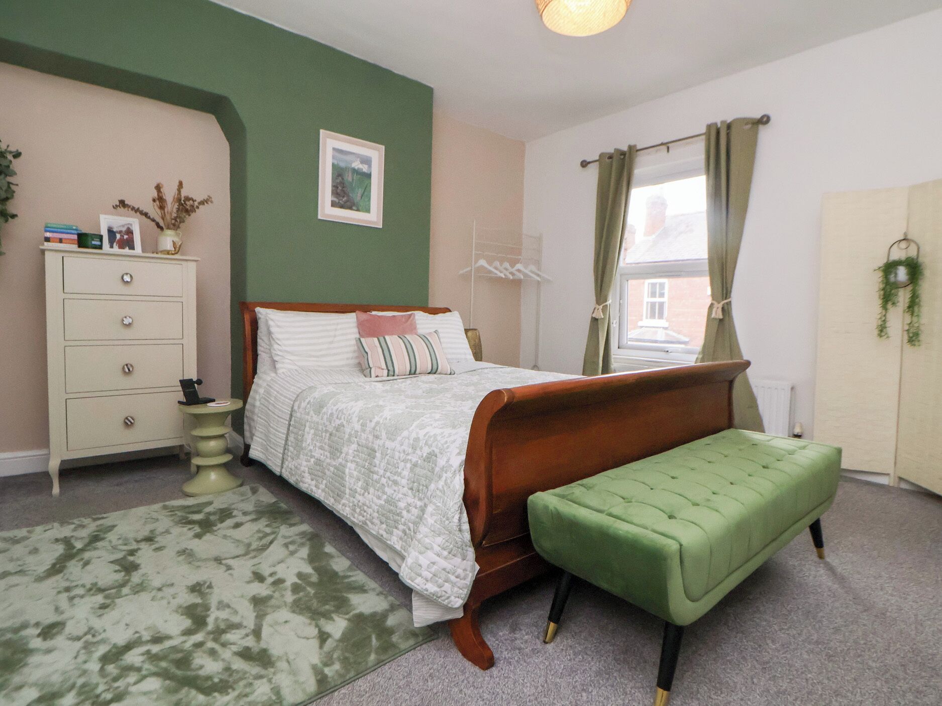 Un dormitorio con cama, cómoda, banco verde, radiador y ventana con cortinas.