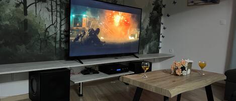 A flat-screen TV on a stand, a wooden coffee table with drinks and flowers, and a wall with a nature-themed mural.