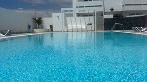 A clear blue swimming pool with lounge chairs and a modern building in the background. - Casahost Dolce Vita Villa Eira (Costa Teguise)