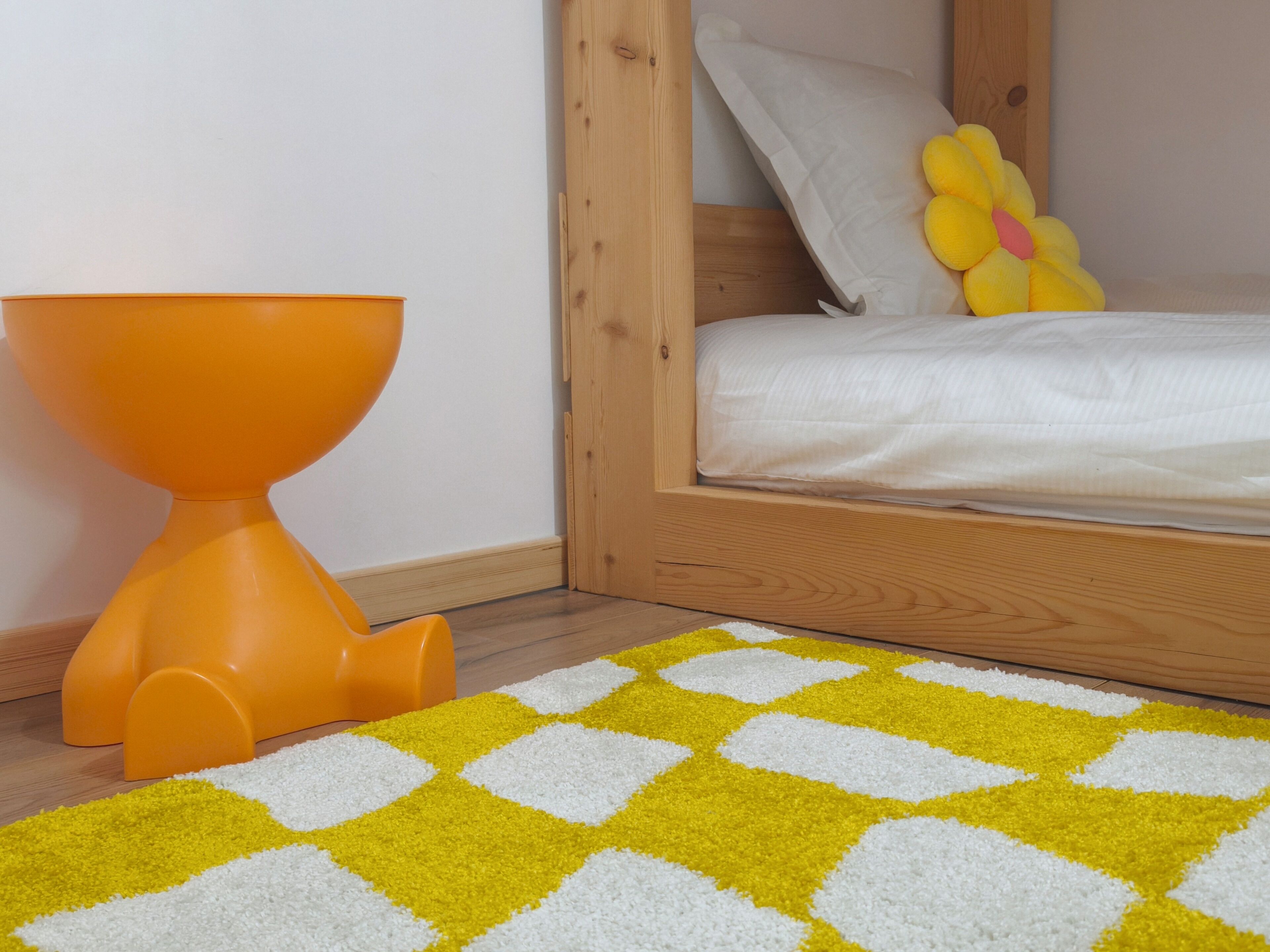 A child's room with a bed, a wooden headboard, a yellow flower pillow, and a yellow and white checkered rug.