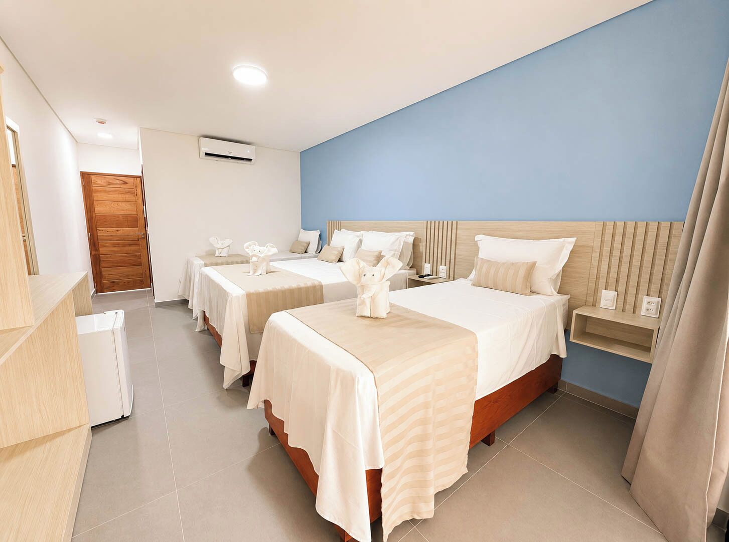 Habitación de hotel con tres camas, cabecera de madera, mesita de noche y estante empotrado.