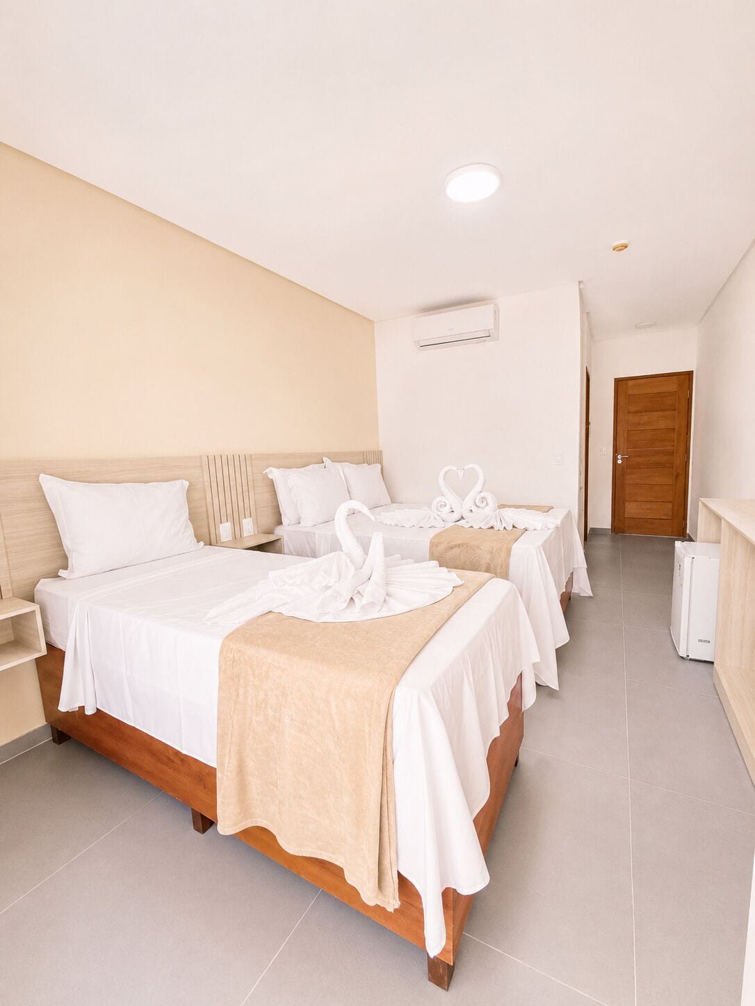 Habitación de hotel con una cama grande, dos camas más pequeñas, un cabecero de madera, una pared blanca, una puerta de madera y un equipo de aire acondicionado blanco.