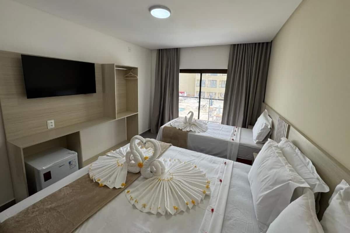 Ein Hotelzimmer mit Flachbildfernseher, einem Bett mit weißer Bettwäsche und dekorativen Schwanenkissen, einem Nachttisch und einem Fenster mit Vorhängen.