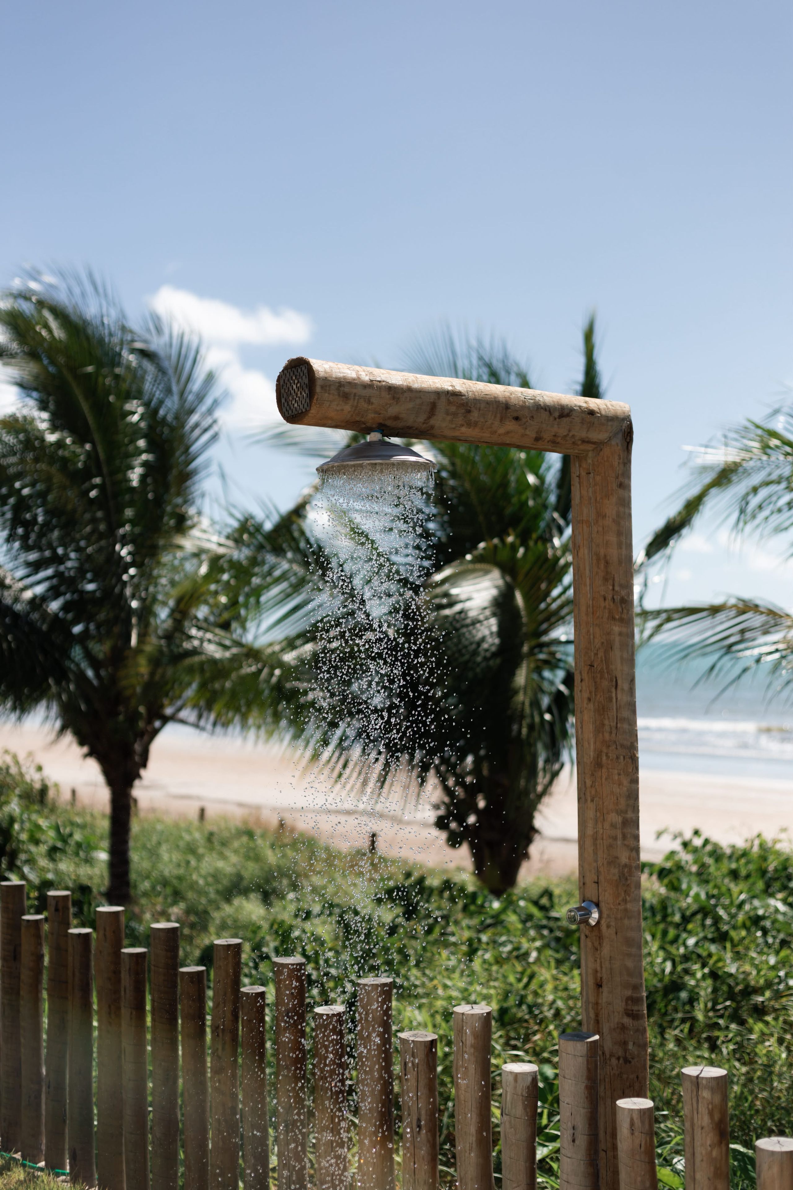 Eine hölzerne Konstruktion mit Wasserspeier, umgeben von Grünpflanzen und Palmen, mit Blick auf einen Strand.