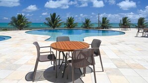 Uma área de piscina com mesa e cadeiras de madeira, cercada por palmeiras e um céu azul claro.