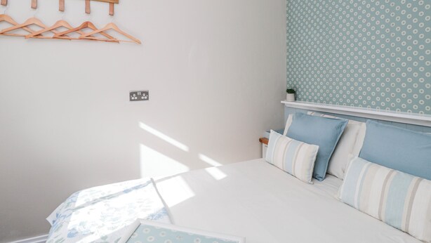 A bedroom with a bed, a wall-mounted light fixture, a wooden hanger rack, and a patterned wall. - Firefly Cottage (Cirencester)