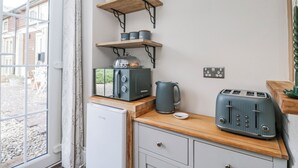 A compact kitchen with a refrigerator, toaster, kettle, and shelves with jars. - Firefly Cottage (Cirencester)