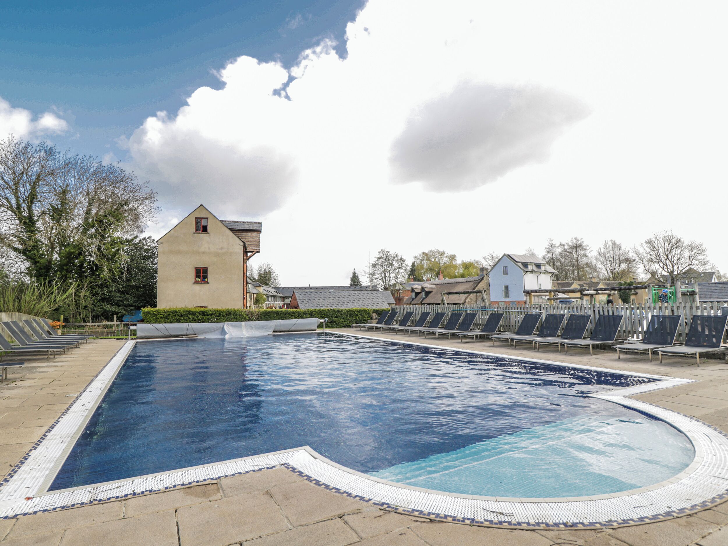 A swimming pool with a tiled edge, surrounded by a fence, and buildings in the background.