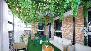Une pièce avec un mur en briques, du gazon synthétique vert, des canapés blancs, des fauteuils en osier et une table basse ronde sur laquelle est posé un vase.