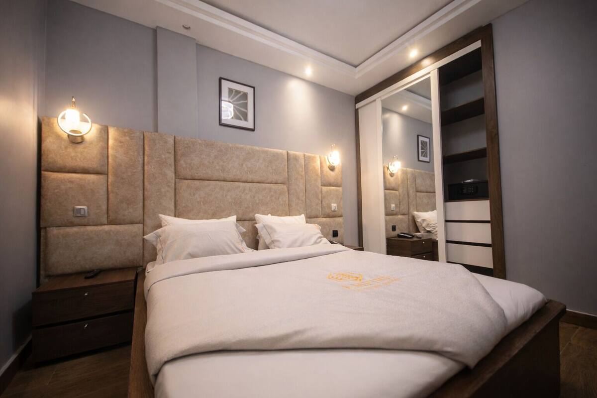 Una habitación de hotel moderna con una cama grande, cabecera con iluminación integrada, mesita de noche y armario con espejo.