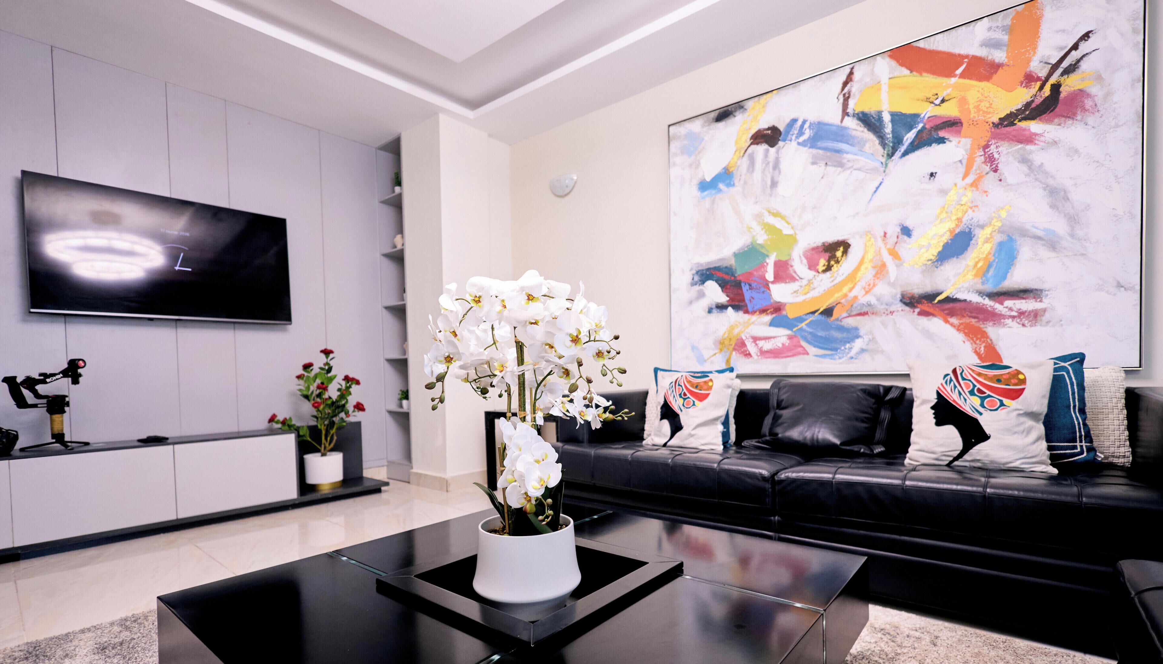 A modern living room with a flat-screen TV, a black leather sofa, a coffee table, and a large abstract painting.