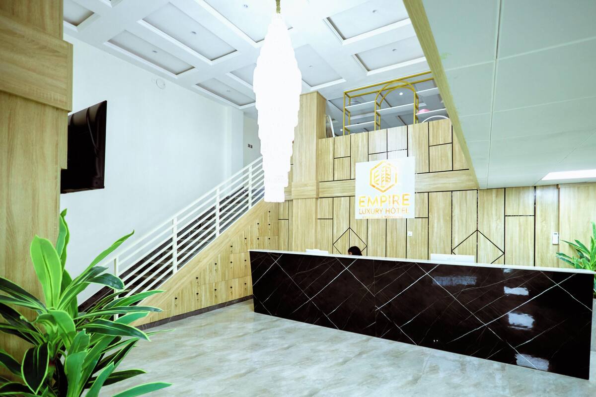 Zona de recepción de hotel con diseño moderno, un mostrador de recepción, una escalera y un cartel que dice 'EMPIRE LUXURY HOTEL'.