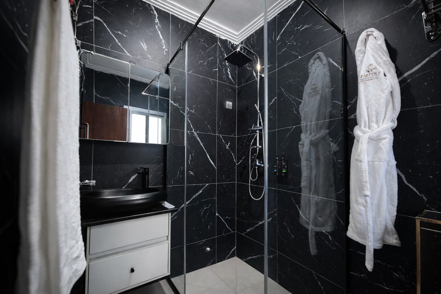 A modern bathroom with black marble tiles, a glass-enclosed shower, a white robe hanging on the wall, and a white sink with a mirror above it.