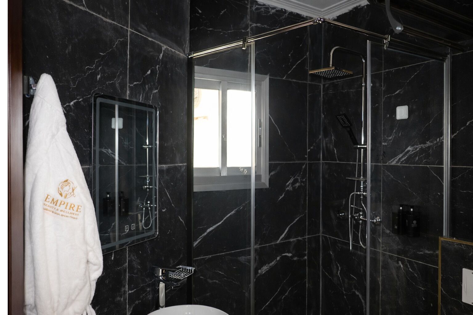Een moderne badkamer met zwart marmeren wanden, een douche met glazen wanden, een witte handdoek en een raam dat natuurlijk licht binnenlaat.