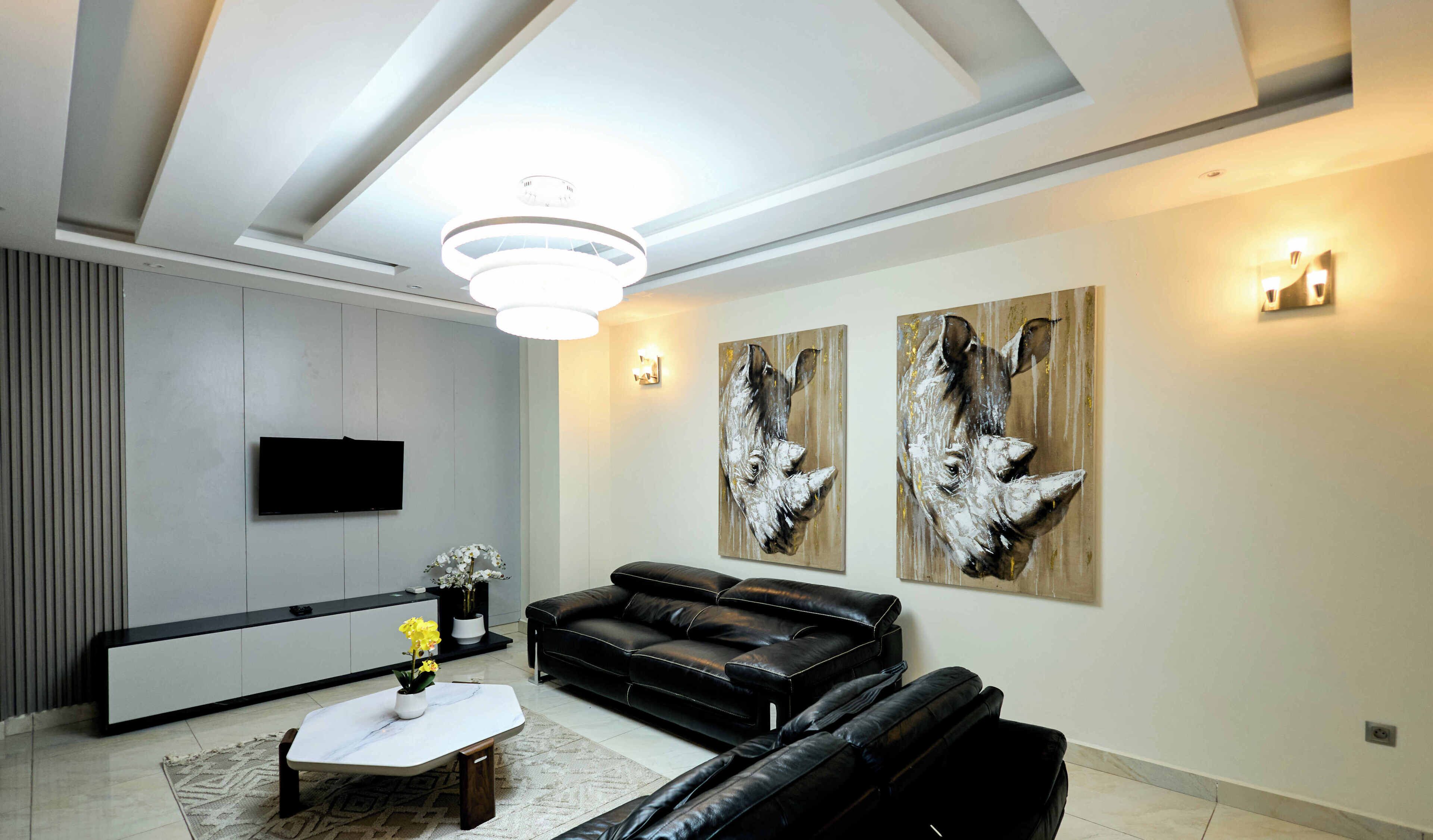 A modern living room with a black leather sofa, a white coffee table, a flat-screen TV, and two abstract paintings on the wall.