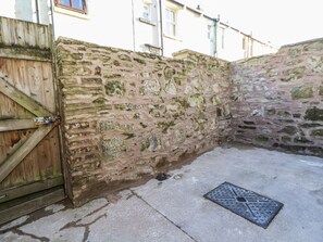 Un muro de piedra con una verja de madera, una tapa de alcantarilla metálica y un suelo de hormigón.