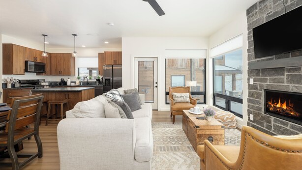 A modern kitchen with a central island, a dining area with wooden chairs, a living room with a sofa and a fireplace, and a large window providing a view of the outdoors. - Base Camp Townhome #106 Slopeside 3 bedroom townhome, views, private balcony (Granby)