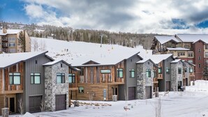 A modern multi-story building with a mix of stone and wood exterior, multiple garage doors, and a ski lift in the background. - Base Camp Townhome #106 Slopeside 3 bedroom townhome, views, private balcony (Granby)