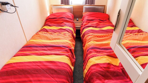 Ein schmales Zimmer mit zwei Betten, jeweils mit gestreifter Bettwäsche, einem Nachttisch und einer Steckdose an der Wand.