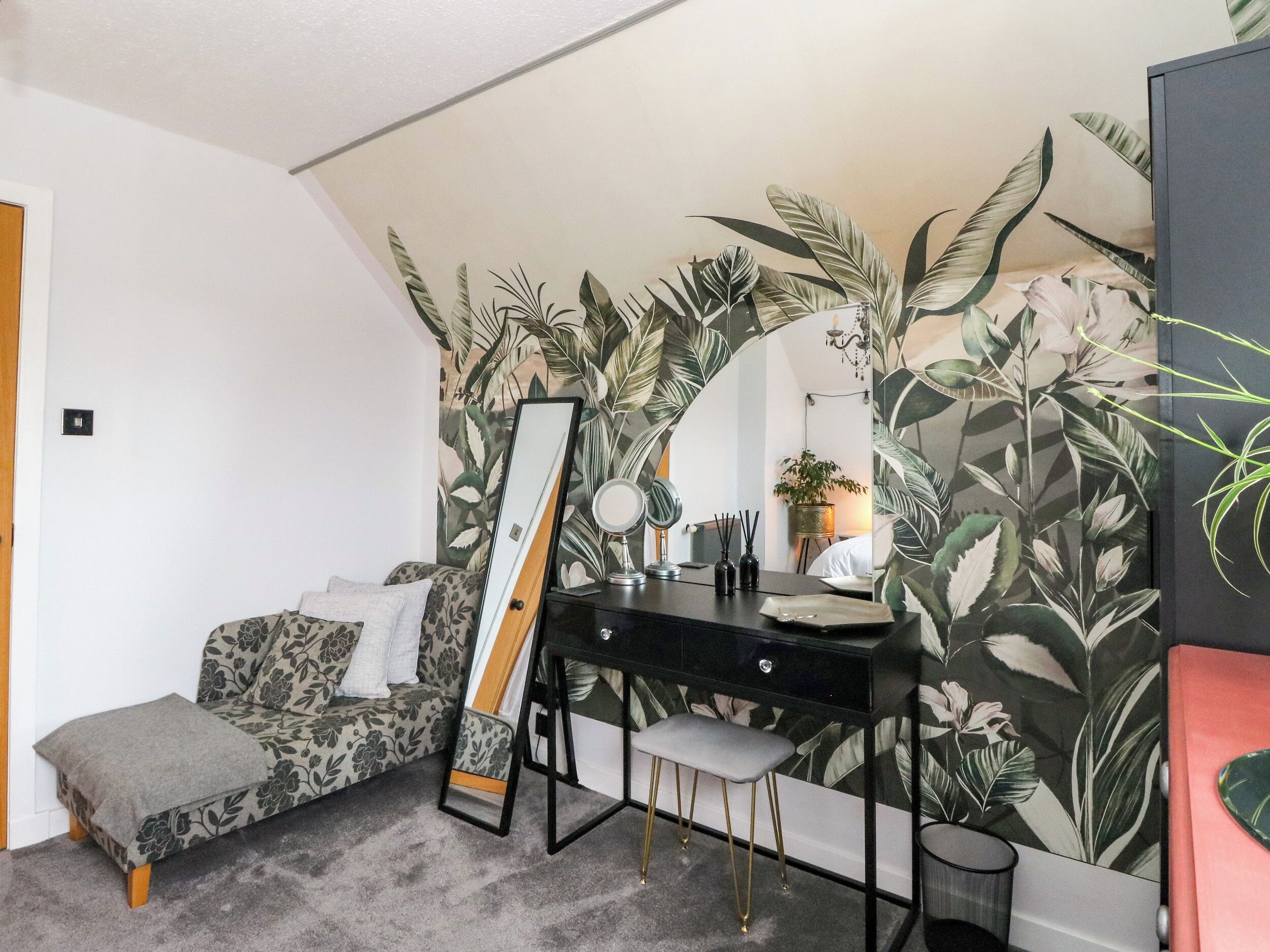 Una habitación con cama, un escritorio con espejo, un taburete pequeño y una pared con motivos de hojas tropicales.