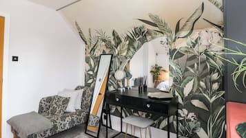 Una habitación con cama, un escritorio con espejo, un taburete pequeño y una pared con motivos de hojas tropicales.