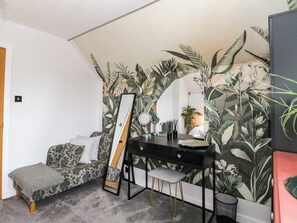 Una habitación con cama, un escritorio con espejo, un taburete pequeño y una pared con motivos de hojas tropicales.