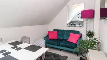 A modern living room with a white ceiling, a teal sofa with pink cushions, a white table with black placemats, a gray furry rug, and a mirror on the wall.