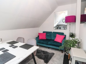 Ein modernes Wohnzimmer mit weißer Decke, einem petrolfarbenen Sofa mit rosa Kissen, einem weißen Tisch mit schwarzen Platzsets, einem grauen, flauschigen Teppich und einem Spiegel an der Wand.