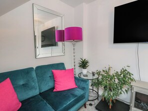 A living room with a teal sofa, a pink cushion, a round table with a potted plant, and a flat-screen TV mounted on the wall. - 3 Linshader (Isle of Lewis)