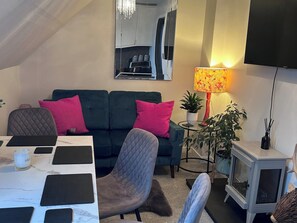 A cozy living room with a sofa, a dining table with black placemats, a small side table with a lamp, a television, and a decorative plant. - 3 Linshader (Isle of Lewis)