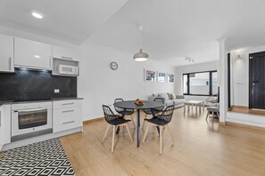 Eine moderne Küche mit weißen Schränken, einer Mikrowelle und einem eingebauten Backofen. Es gibt einen Essbereich mit einem runden Tisch und Stühlen sowie ein Wohnzimmer mit einer Couch und einem Fenster.