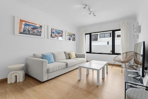 Ein modernes Wohnzimmer mit einer Couch, einem Couchtisch und einem Fernseher. An den Wänden hängen Bilderrahmen und ein Fenster mit Vorhängen.