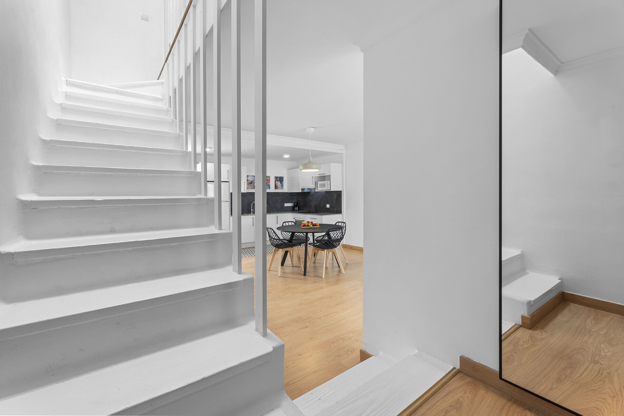 A modern interior with a staircase, a dining area with a round table and chairs, and a kitchen in the background.