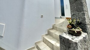 Un edificio moderno blanco con una escalera que lleva a un balcón, un pilar de piedra con plantas y un cielo azul despejado.