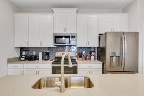 A modern kitchen with white cabinets, a stainless steel sink, a microwave, a stove, and a refrigerator. - 8-Min Disney Villa with Resort & Pool (Kissimmee)