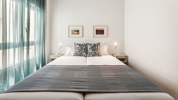 A hotel room with a large bed, two bedside tables, a window with curtains, and two framed pictures on the wall. - Dragos Suites 202 (Arrecife)