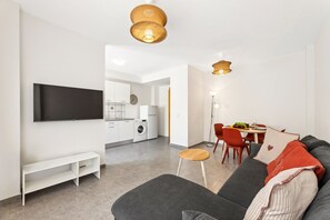 A modern living room with a flat-screen TV, a grey sofa, a small wooden stool, and a dining area with a table and chairs. - Dragos Suites 202 (Arrecife)