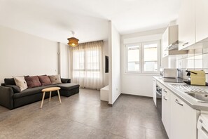 A modern living room with a sofa, a small wooden table, and a kitchen area with white cabinets and a sink. - Dragos Suites 202 (Arrecife)